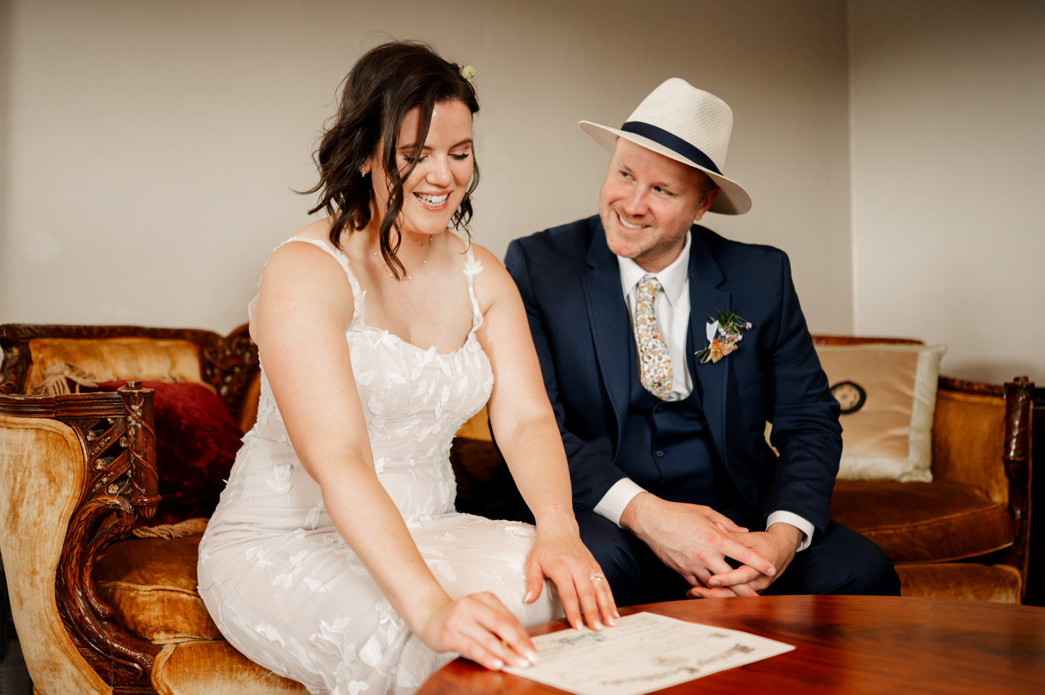 At Rossi Farms, a bride and groom sit on a vintage couch, smiling warmly. The bride looks at a document on a wooden table in front of them.