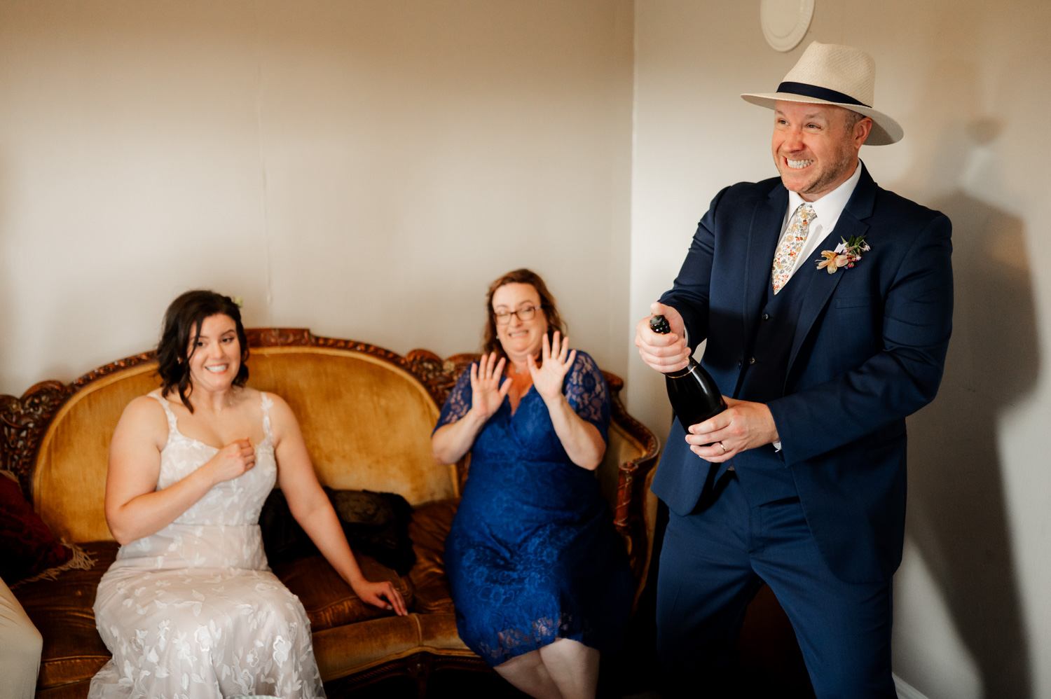 A man in a suit opens a champagne bottle with a smile; two women on a vintage sofa react with joy in an elegantly decorated room, conveying the celebration at Rossi Farms wedding.