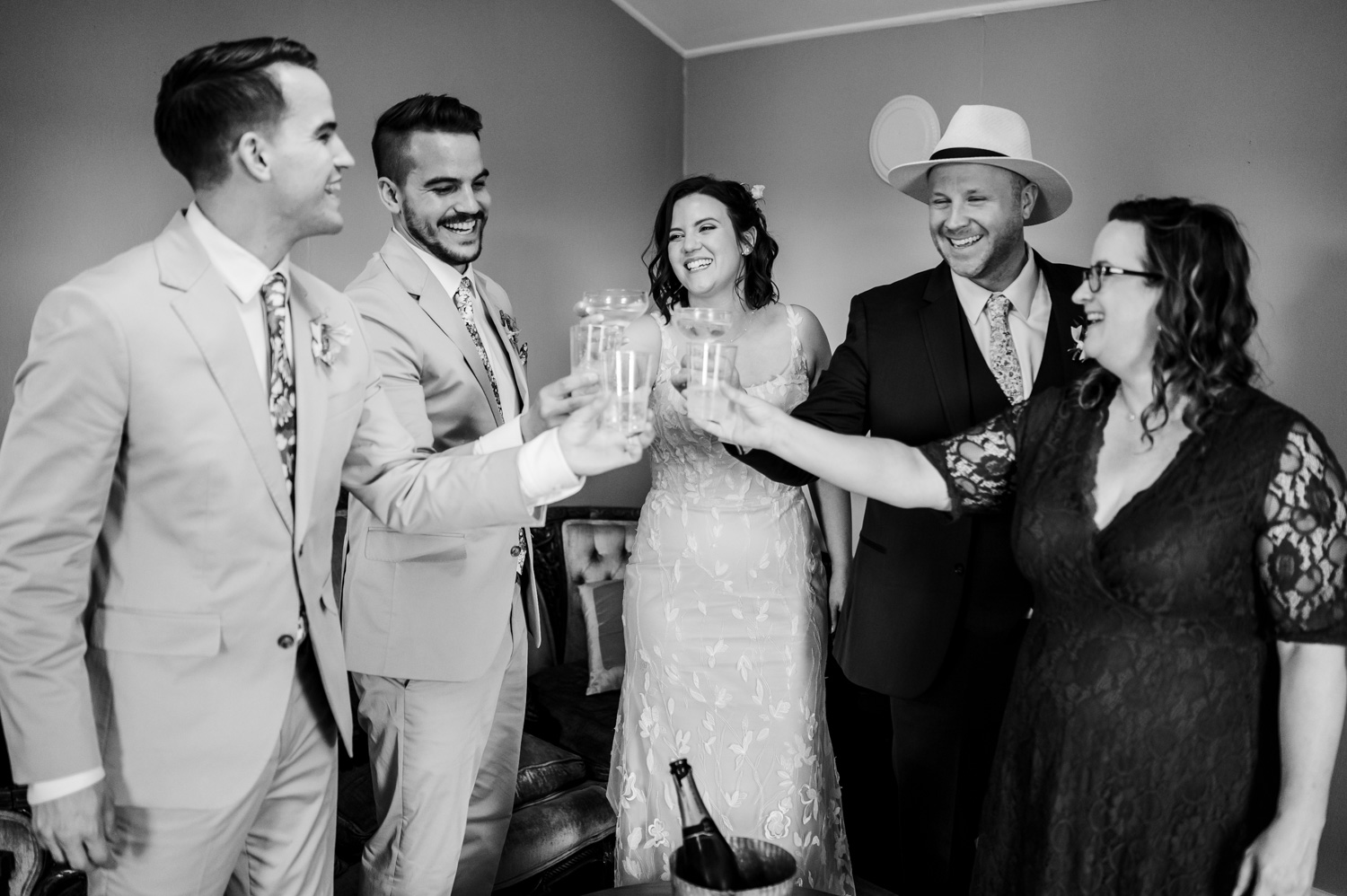 In a cheerful and relaxed indoor setting, five people in formal attire clink their glasses. The moment is filled with joy, with everyone smiling and enjoying the celebration. The elegant backdrop complements the scene, adding to the festive atmosphere.