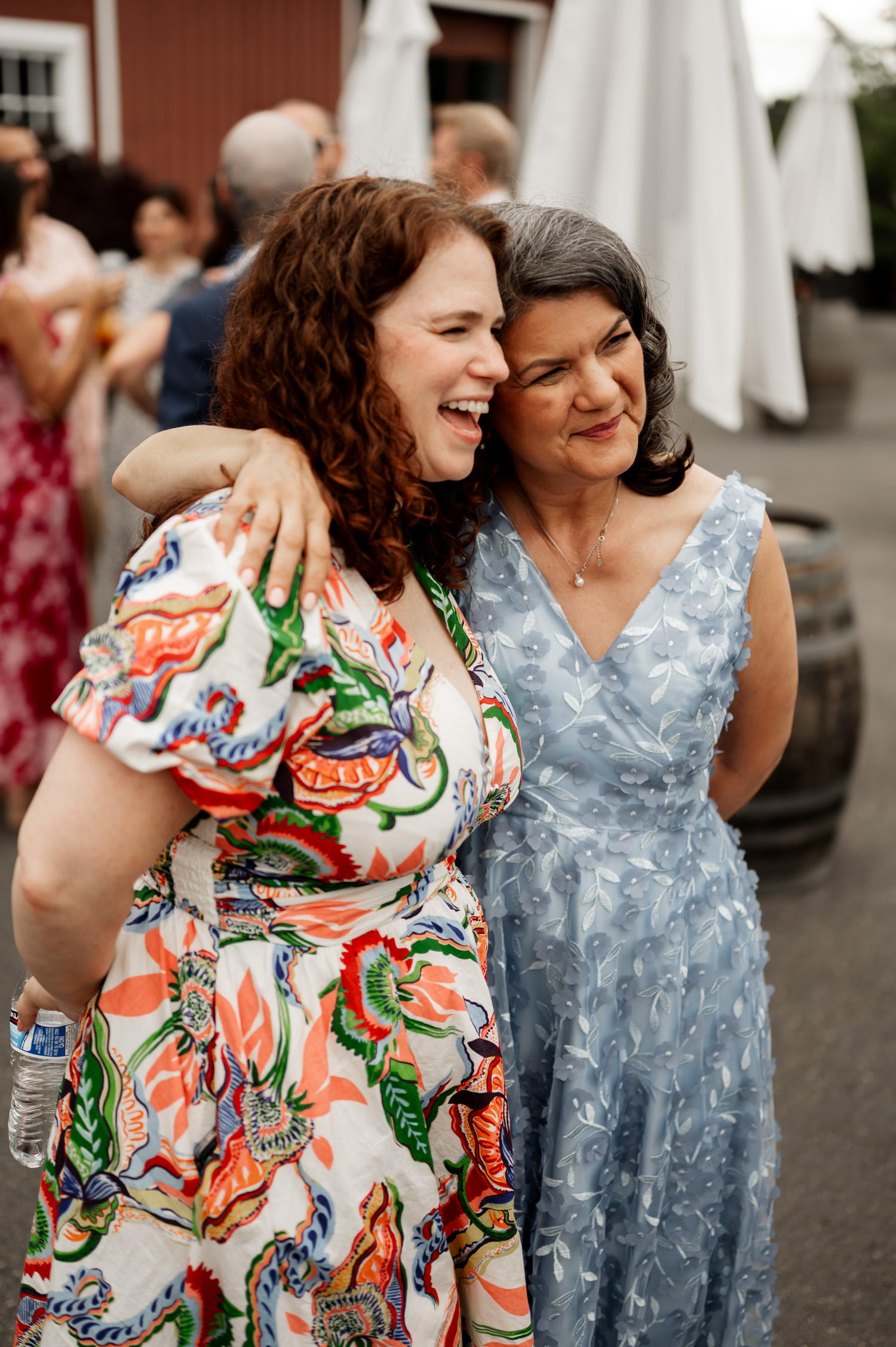 Two women smiling and embracing outdoors, one in a floral dress and the other in a light blue dress, at Rossi Farms.