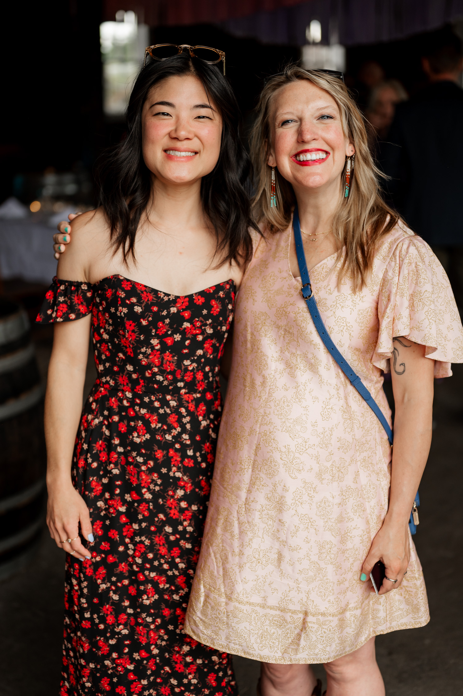 Two joyful women standing side by side indoors, one wearing a floral outfit and the other in a light patterned dress, capturing the warmth and elegance of Rossi Farms wedding photos.