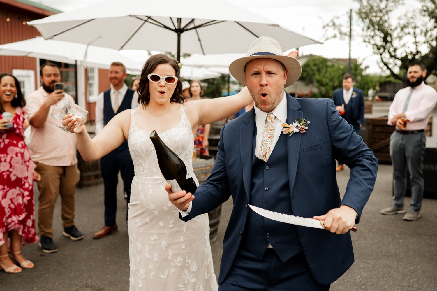 At Rossi Farms, the groom uses a saber to open a champagne bottle outdoors, while the bride stands beside him and guests cheer in the background.