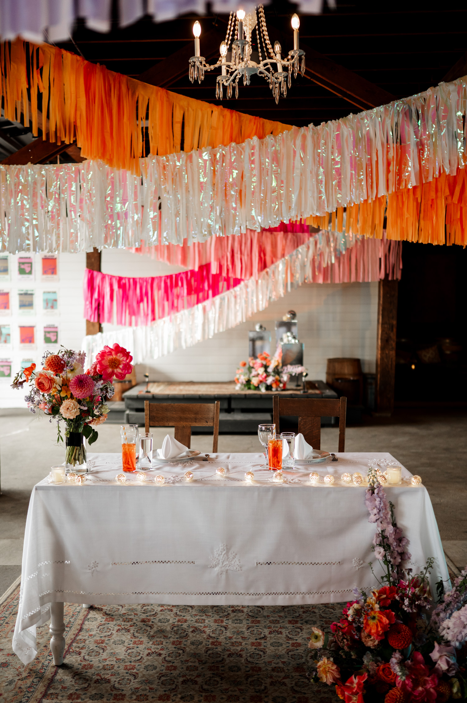 A wedding photo from Rossi Farms shows a table with floral centerpieces, colorful hanging streamers above, and two orange drinks on a clean white tablecloth.