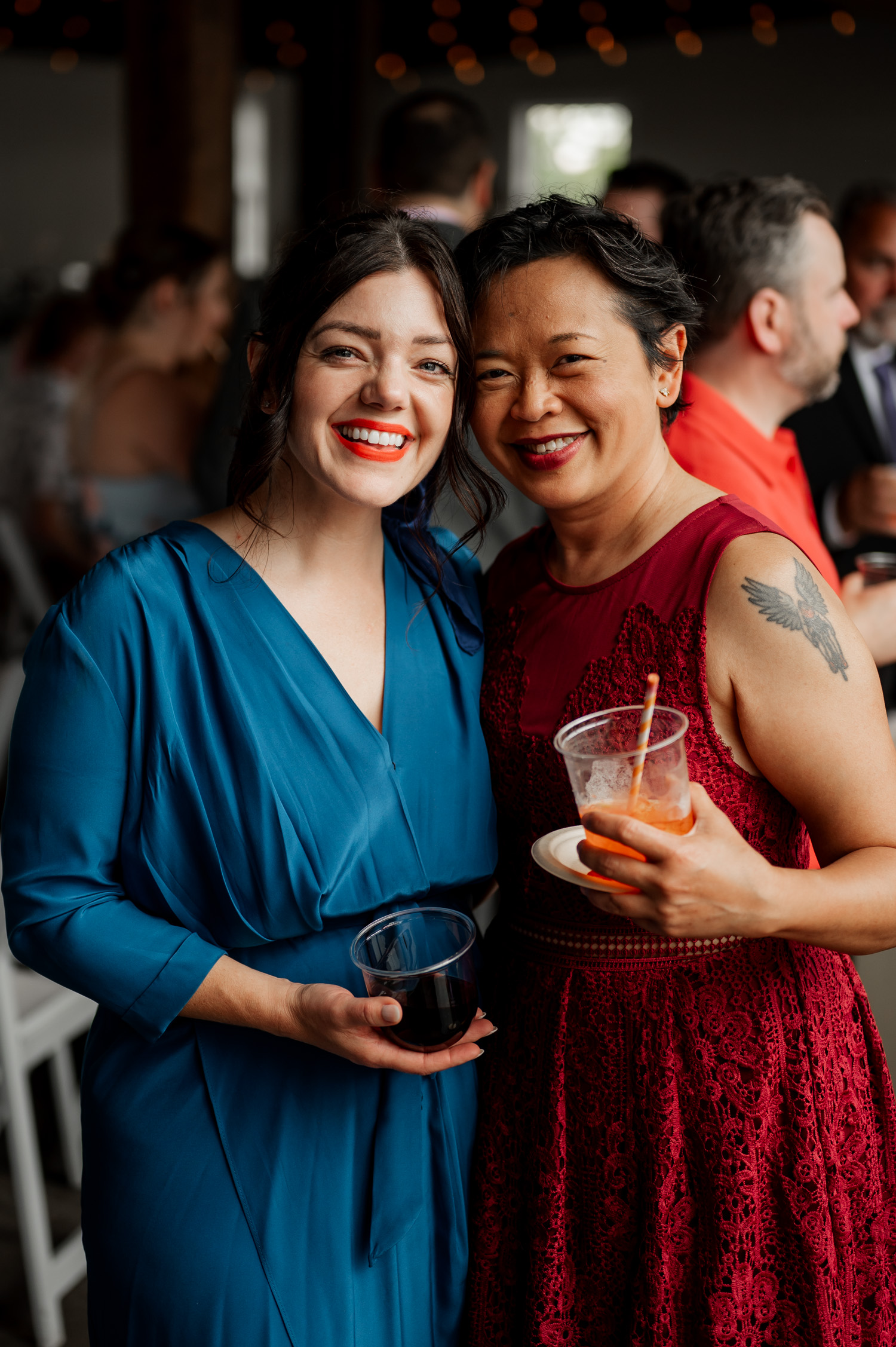 Two women smiling at a party, one wearing a blue dress and the other in a red lace dress holding a drink, enjoying joyful moments.
