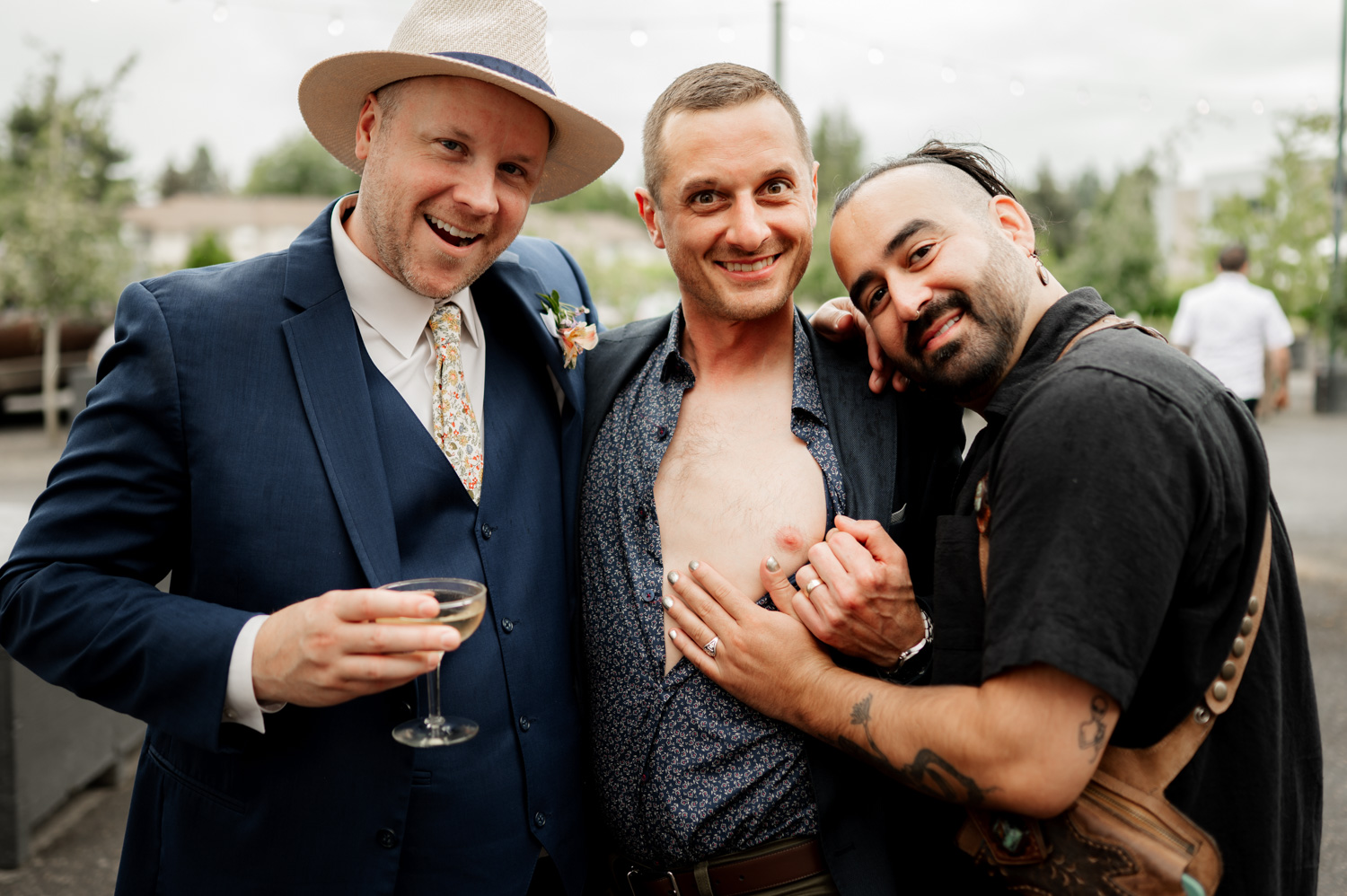 Three men share a joyful moment outside Rossi Farms. One man, wearing a suit, sips a drink as another, dressed in an open shirt, stands beside him. The third man wraps him in a warm embrace, all three smiling brightly.