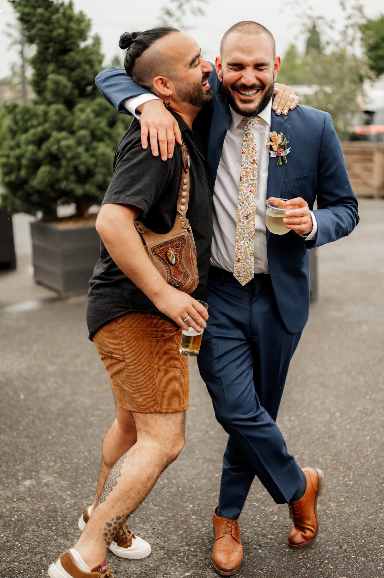 Two men walking outdoors, one wearing a suit and the other in casual attire, both holding drinks and laughing. In the background are wedding decorations from Rossi Farms.