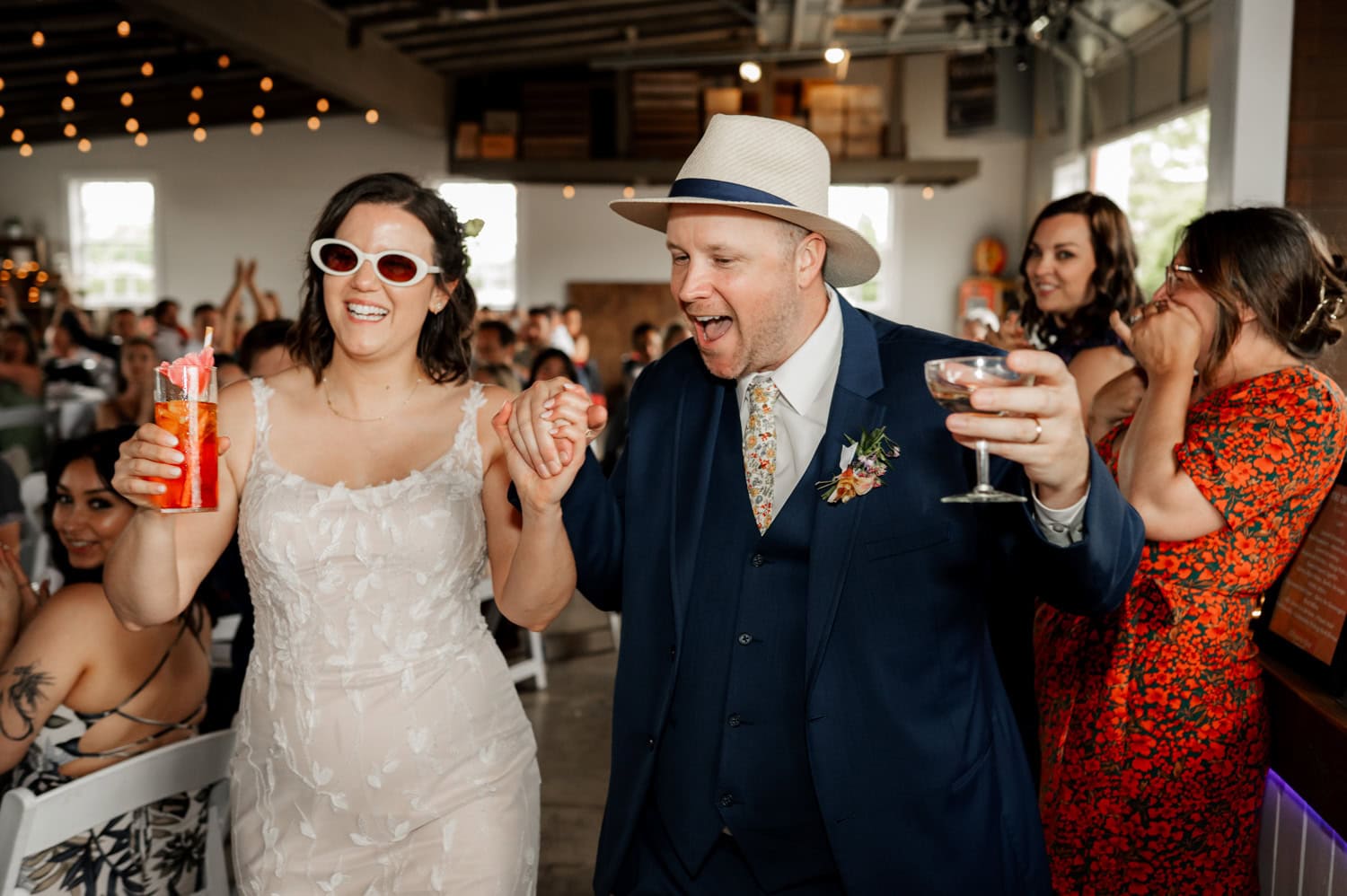 A joyful bride and groom enter the reception at Rossi Farms, holding drinks in one hand and each other’s hand in the other, as cheering guests surround them.