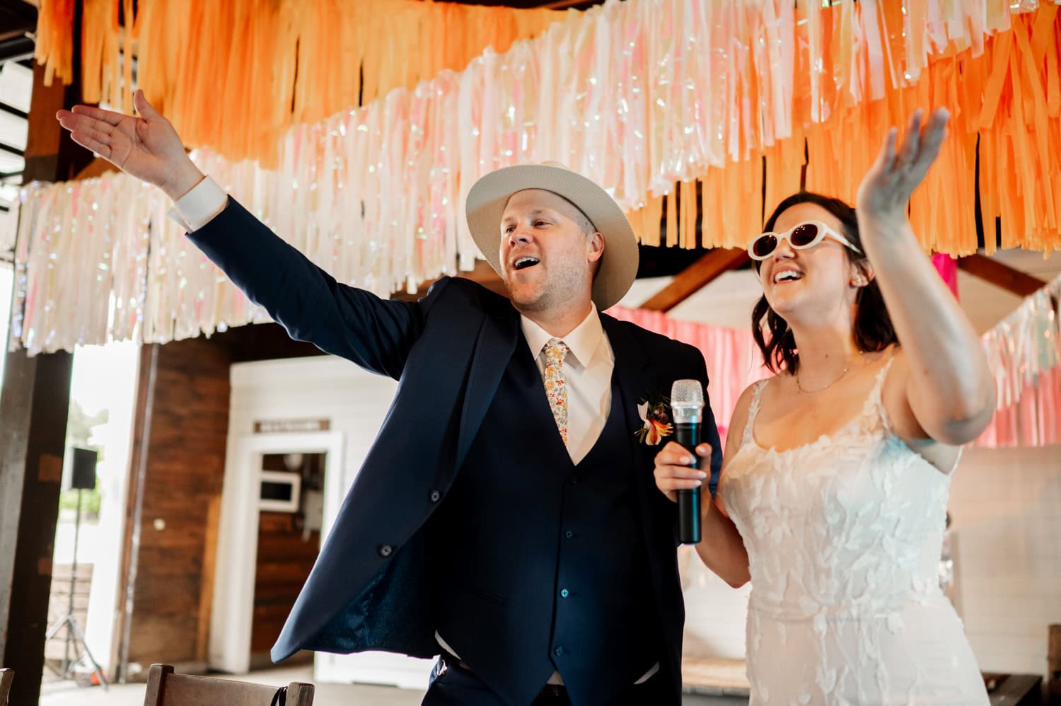A joyful couple in formal attire sings into a microphone, surrounded by colorful streamers at a lively indoor event.