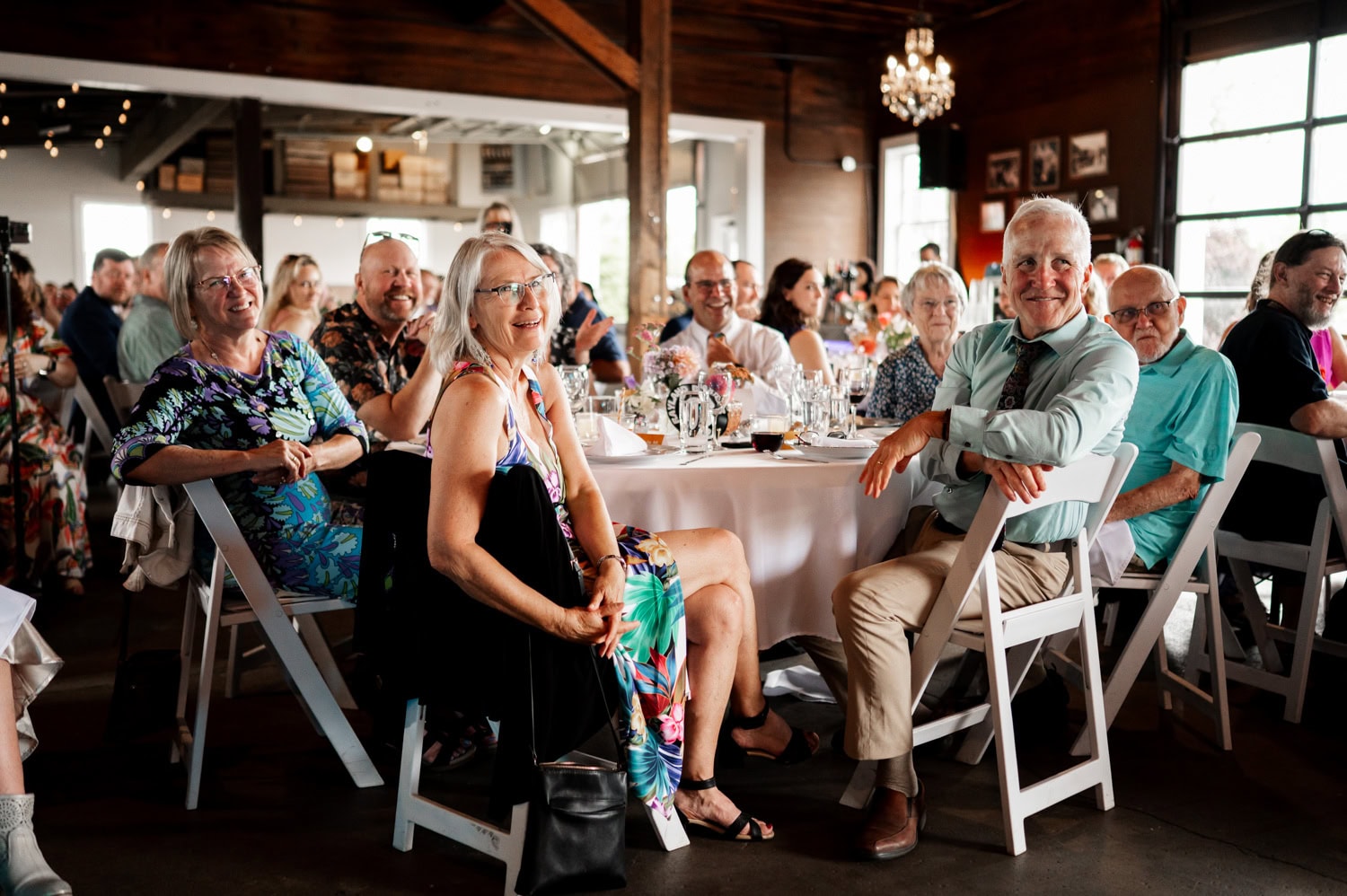 A group of people seated at round tables in a rustic venue, smiling and looking towards the camera during a wedding celebration at Rossi Farms.