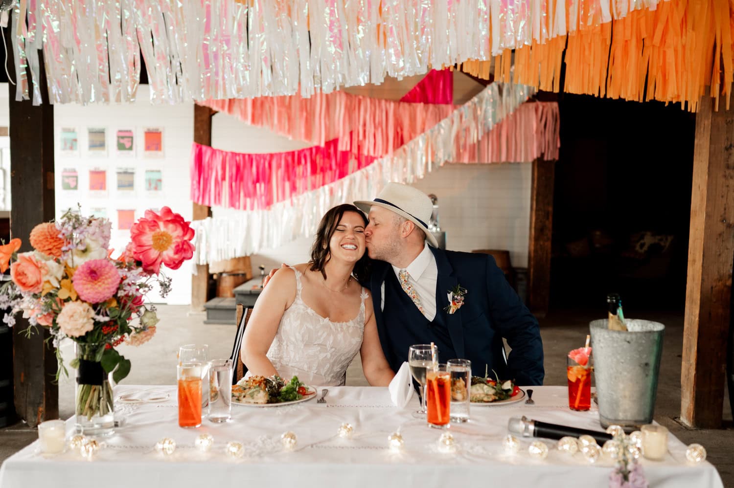 The bride and groom sit at a beautifully adorned wedding table, surrounded by vibrant paper streamers and flowers. The moment is captured perfectly in stunning Rossi Farms wedding photos.