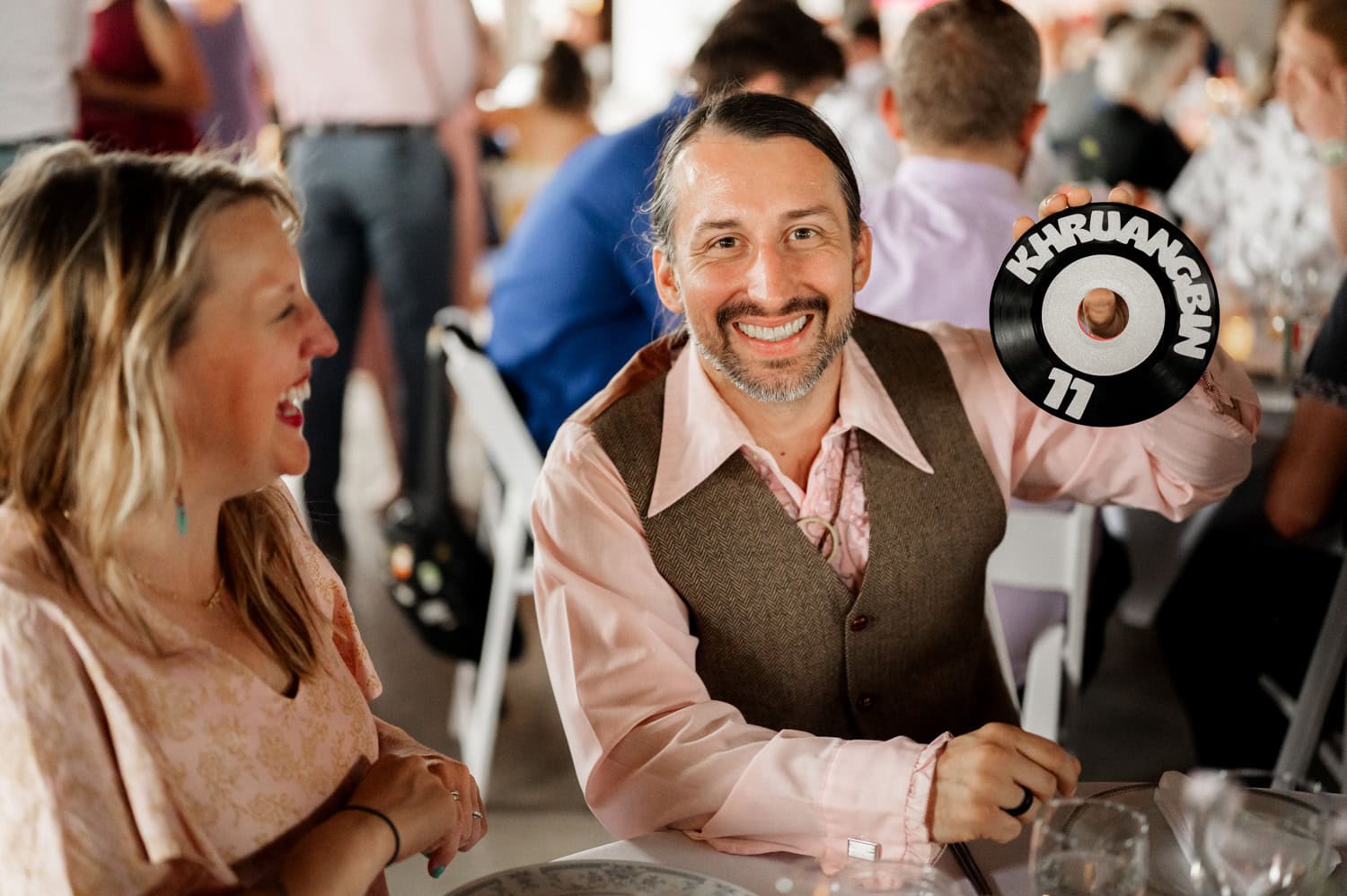 A man smiles while holding a vinyl record with Khruangbin printed on it, sitting beside a woman who is laughing at a Rossi Farms wedding.