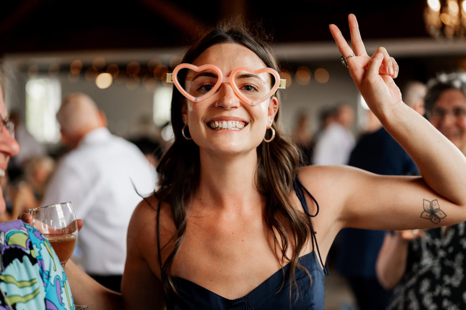 A woman at an indoor event wears heart-shaped glasses and flashes a peace sign with a beaming smile.