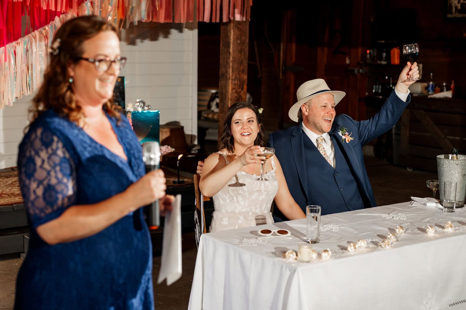 At Rossi Farms, a woman in blue gives a speech while a seated couple in wedding attire raises their glasses for a toast.