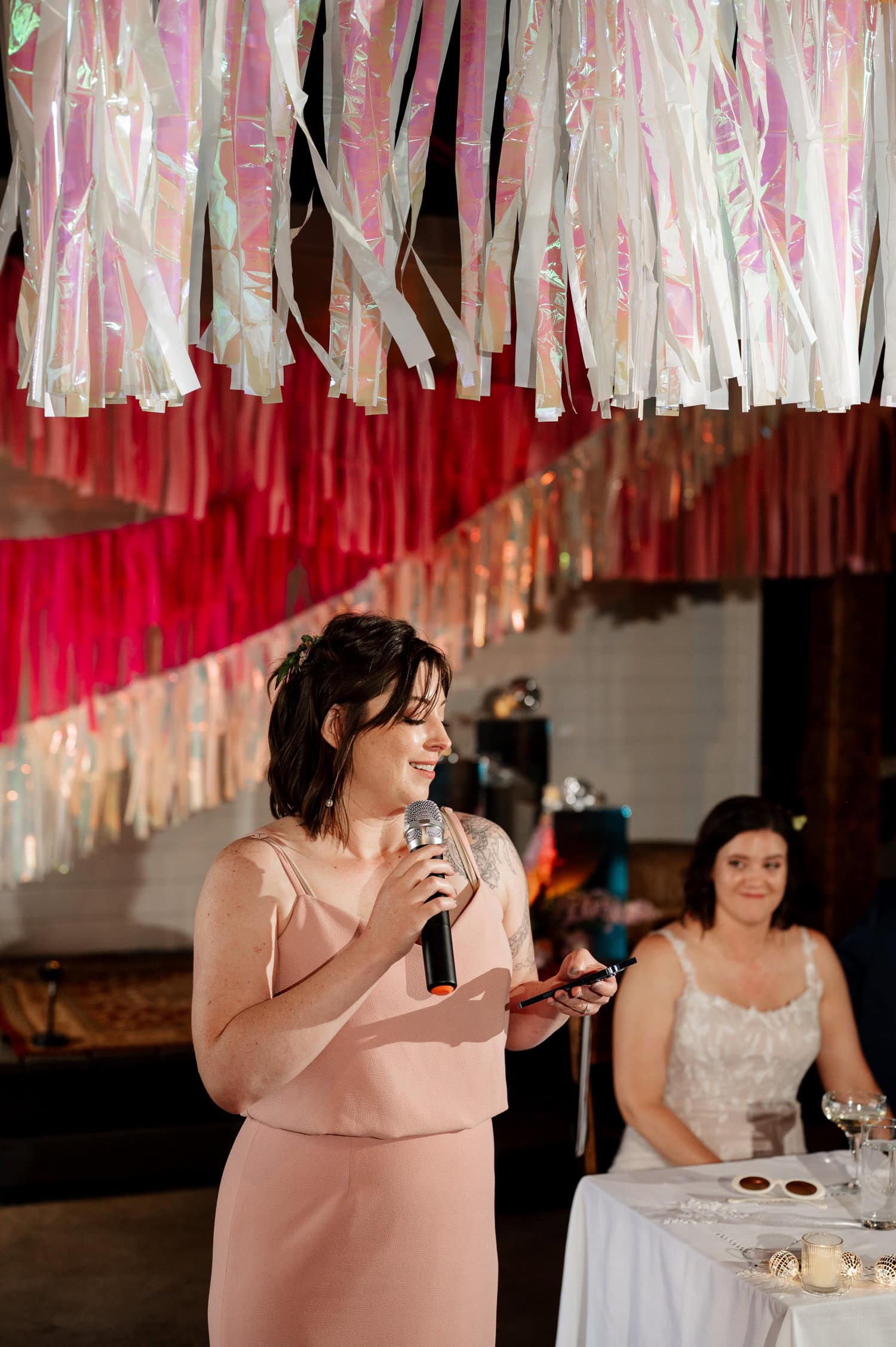 At Rossi Farms, a woman in a peach dress gives a speech with a microphone at the wedding. Colorful decorations hang overhead, adding to the vibrant atmosphere captured in the photos.