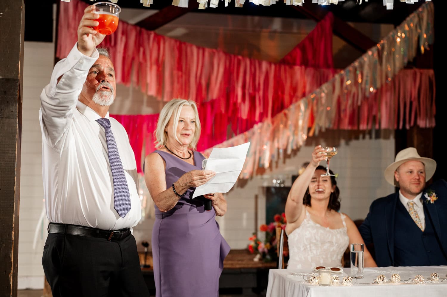 People raising their glasses in celebration at a Rossi Farms wedding, with colorful decorations in the background.