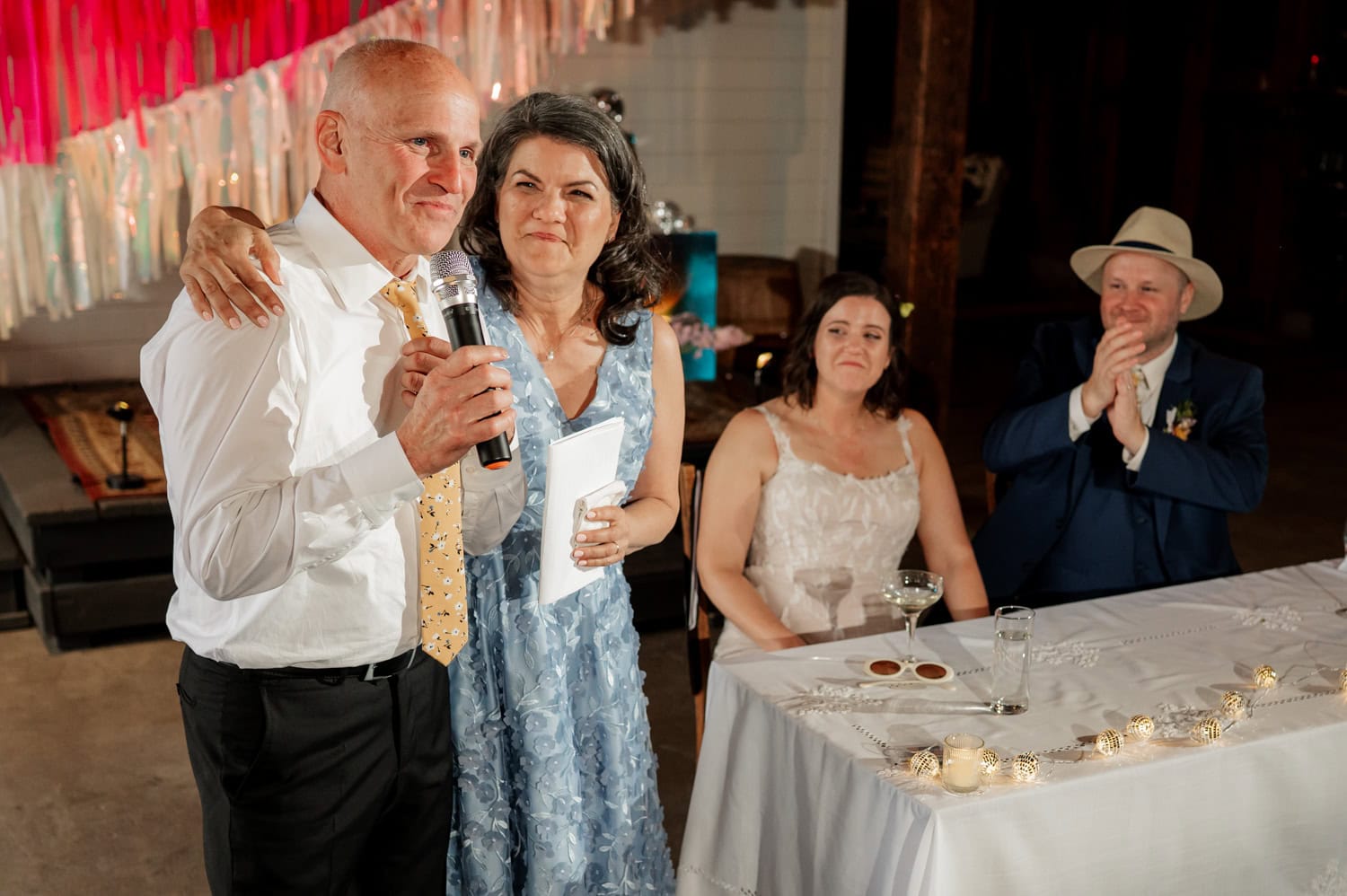 At a Rossi Farms wedding, a man holding a microphone and a woman in a blue dress stand beside the seated couple, capturing precious moments with smiles and warm expressions.