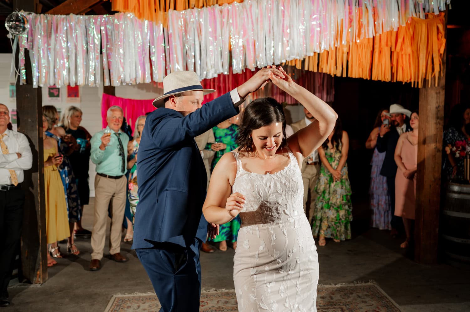 A couple twirls gracefully at a festive event at Rossi Farms, with colorful streamers fluttering overhead and guests watching in the background amidst perfect wedding photo settings.