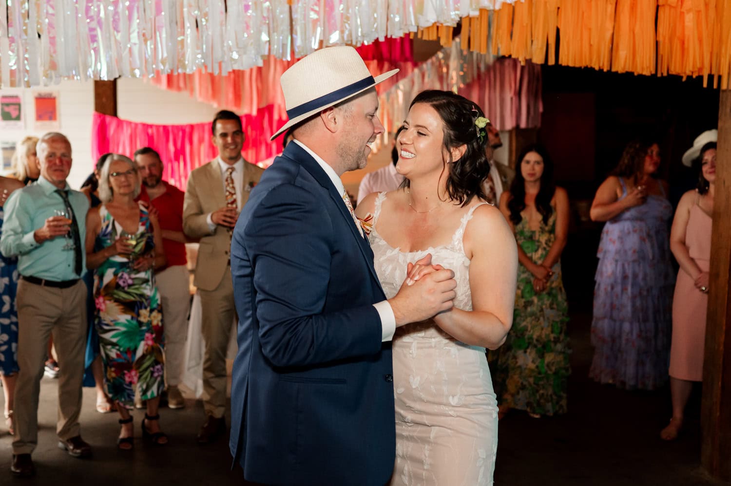The bride and groom wearing hats dance joyfully under colorful streamers at Rossi Farms, surrounded by smiling guests.