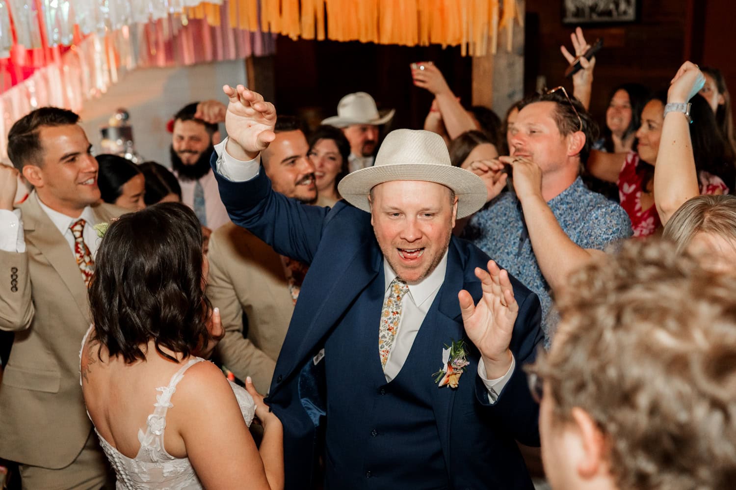 A man wearing a hat dances with joy amidst a crowd and vibrant streamers at a Rossi Farms wedding.