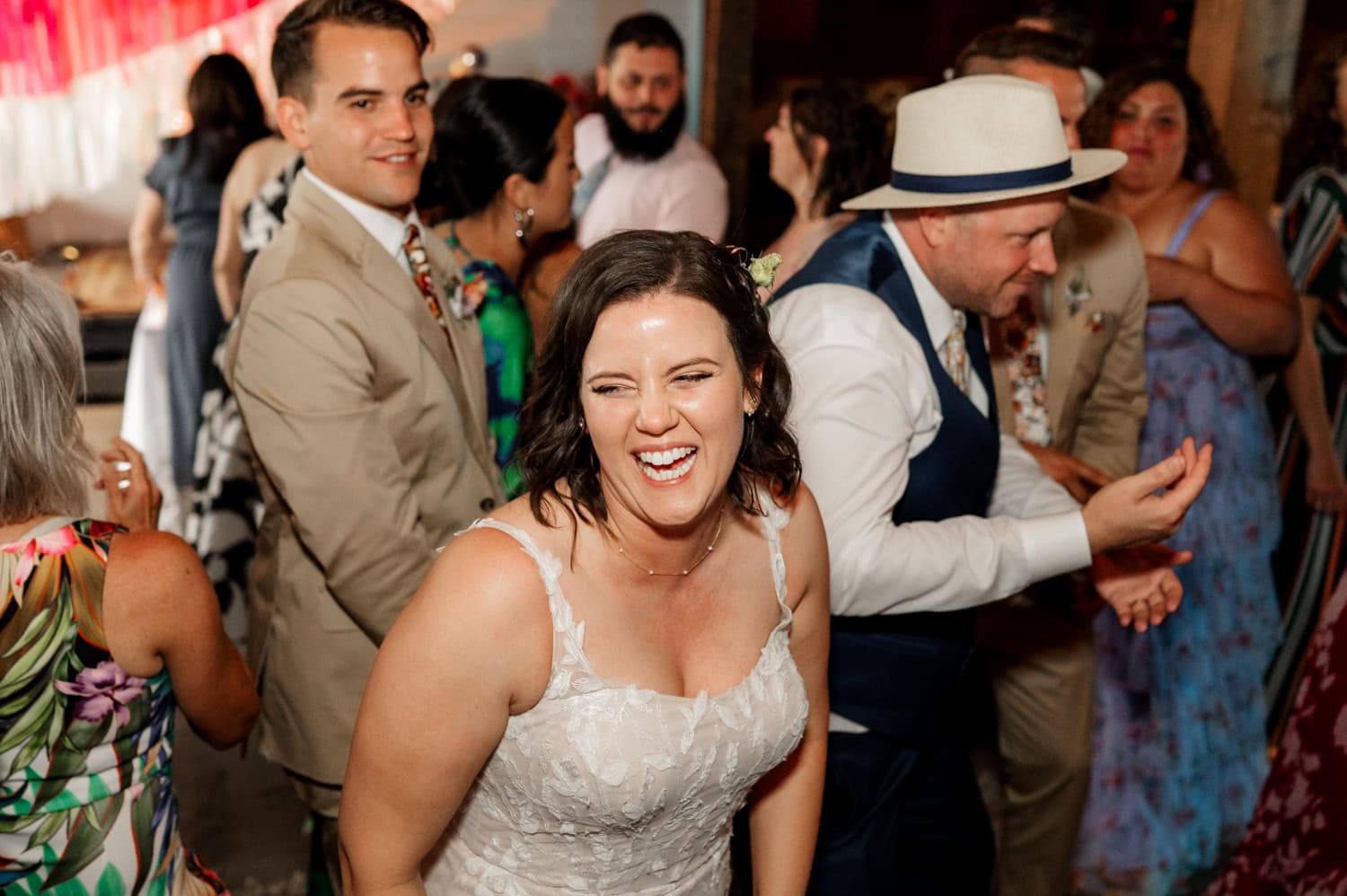 A woman in a white dress is laughing and dancing among guests at a lively indoor celebration, conveying joy and excitement.