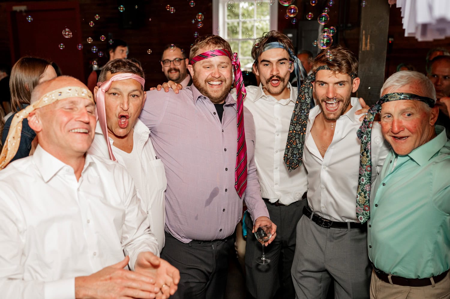 A group of men at a party with ties wrapped around their heads, smiling and posing among bubbles.