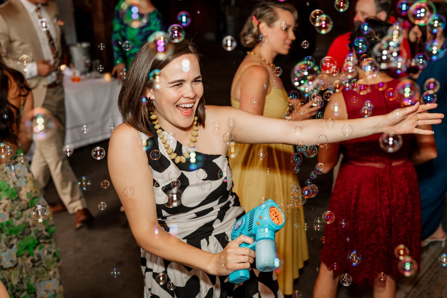 At a lively party, a woman gleefully operates a bubble machine, releasing bubbles into the air. She is surrounded by guests wearing colorful clothing, with a backdrop featuring wedding photos from Rossi Farms.