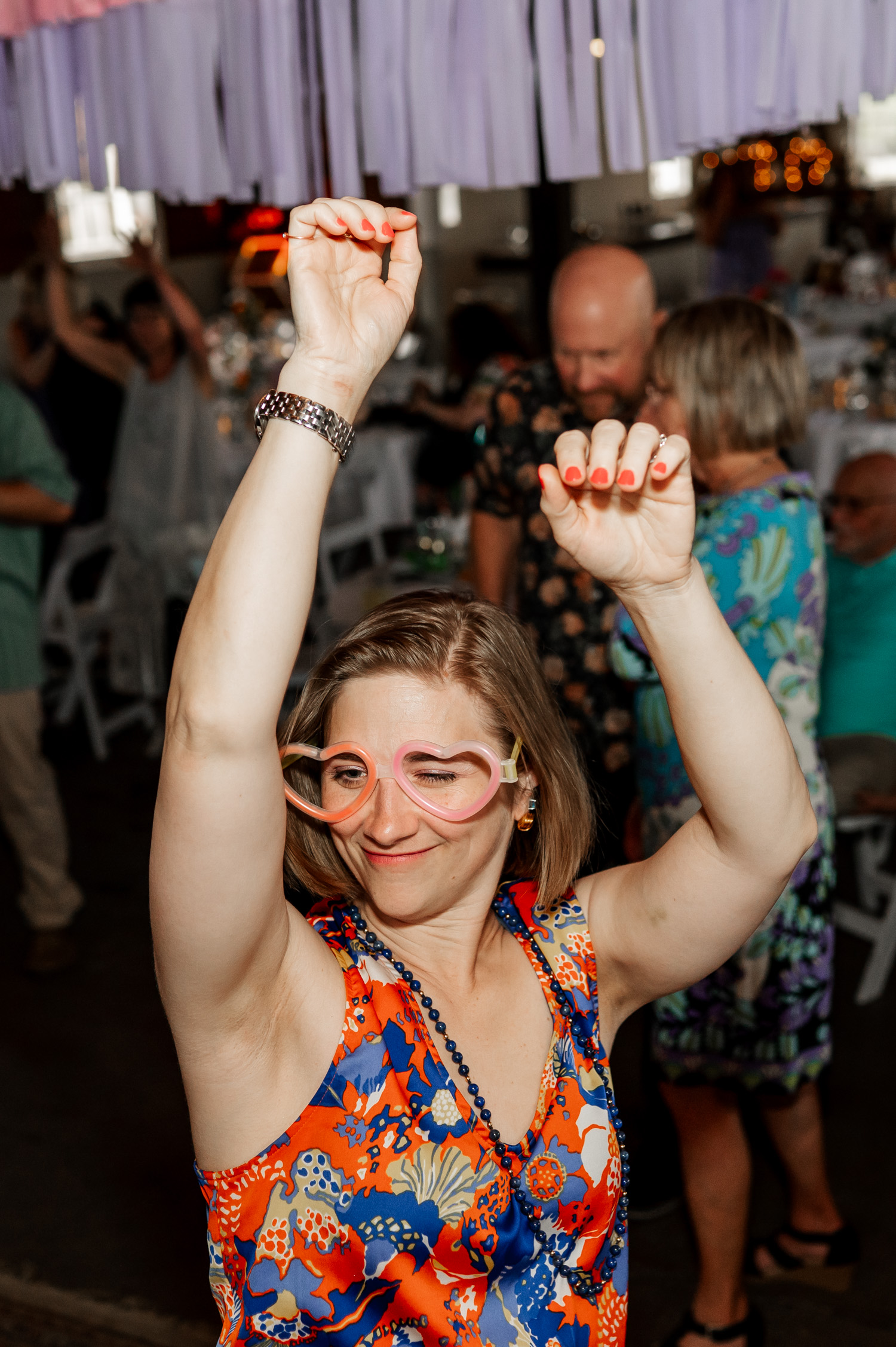 A woman in colorful attire dances energetically at a lively party, surrounded by cheerful guests. Her heart-shaped glasses catch the light as she moves.