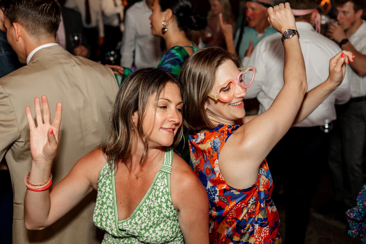Two women in colorful dresses dance joyfully at a lively wedding at Rossi Farms, with other attendees celebrating in the background.