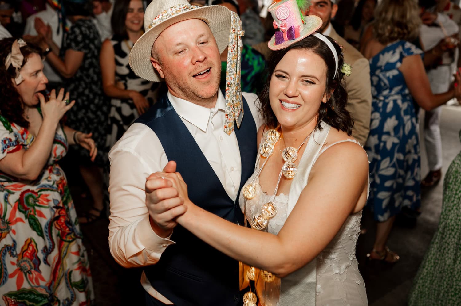 A couple dances joyfully at a lively party, surrounded by guests in colorful outfits and hats, captured beautifully in Rossi Farms wedding photos.
