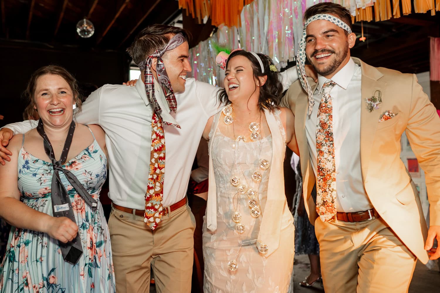 Four people, two women and two men with ties on their heads, are smiling and dancing joyfully at the festive Rossi Farms wedding event.