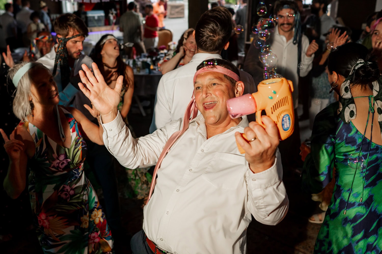A joyful party scene with guests dancing and a man in a white shirt holding a bubble gun, smiling widely at Rossi Farms wedding.