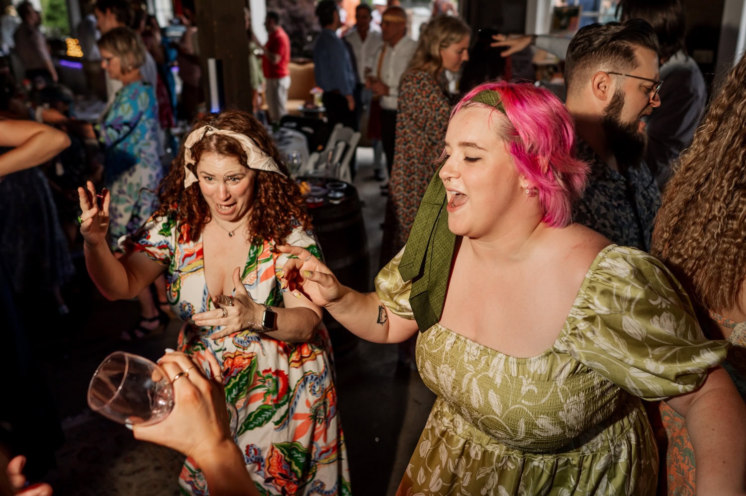 Two women dancing energetically indoors at a Rossi Farms wedding, surrounded by guests; one has pink hair and the other is wearing a bandana.