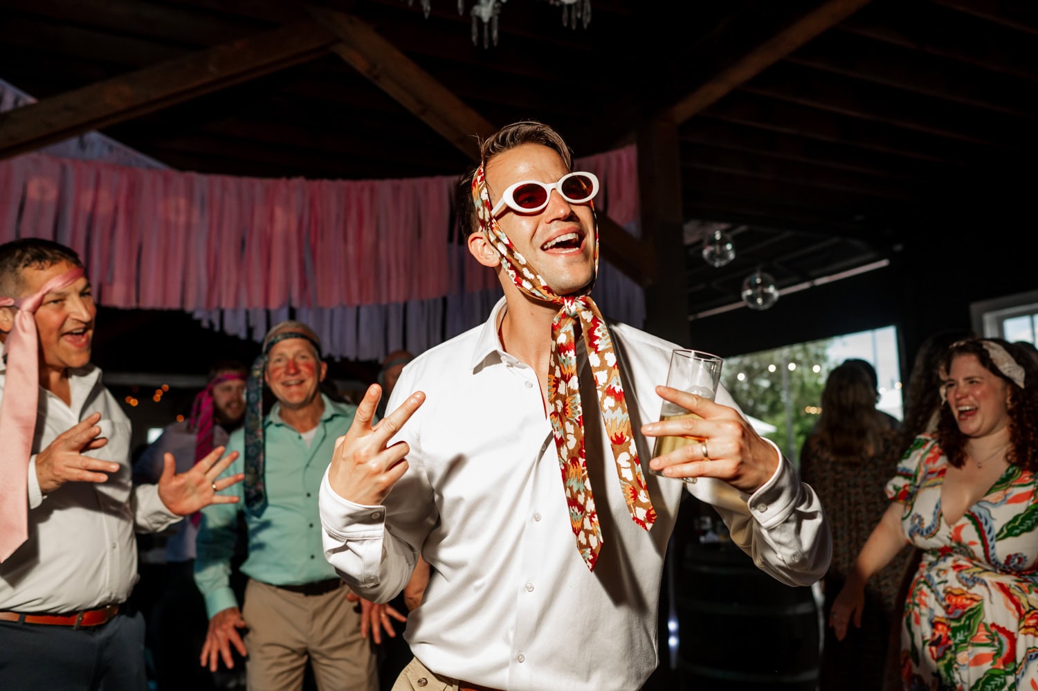 A person wearing sunglasses and a scarf dances joyfully while holding a drink at a lively indoor event. People in the background are smiling and enjoying themselves, contributing to an energetic atmosphere.