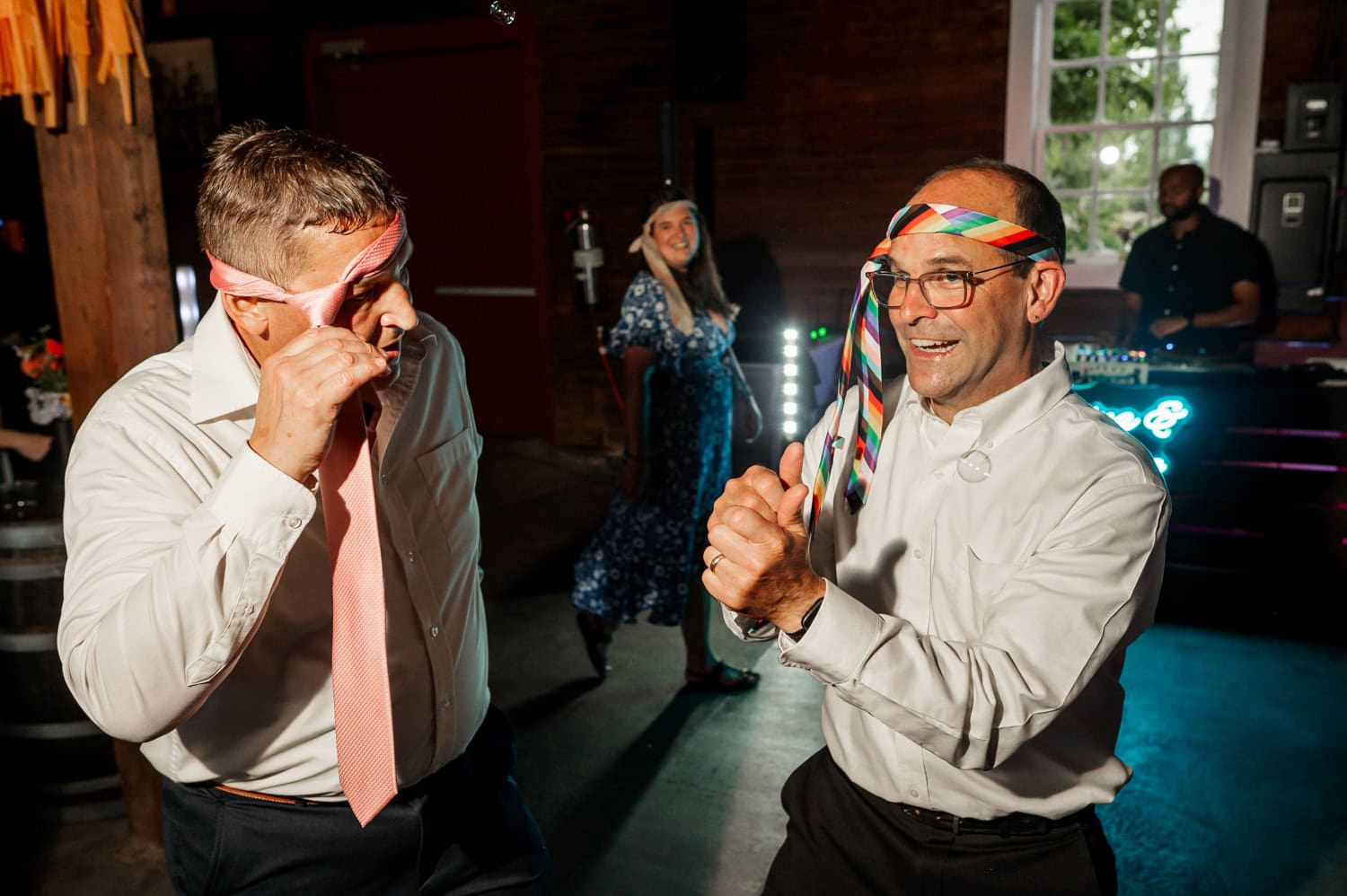 Two men with ties wrapped around their heads dance energetically at a lively party, while a woman and a DJ are visible in the background adding to the festive atmosphere.