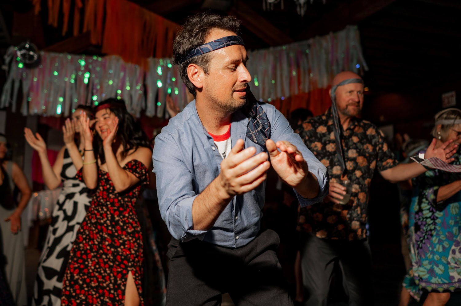 A man dances energetically at a lively celebration, with others surrounding him, clapping and smiling. The scene captures a joyful atmosphere filled with happiness and excitement.