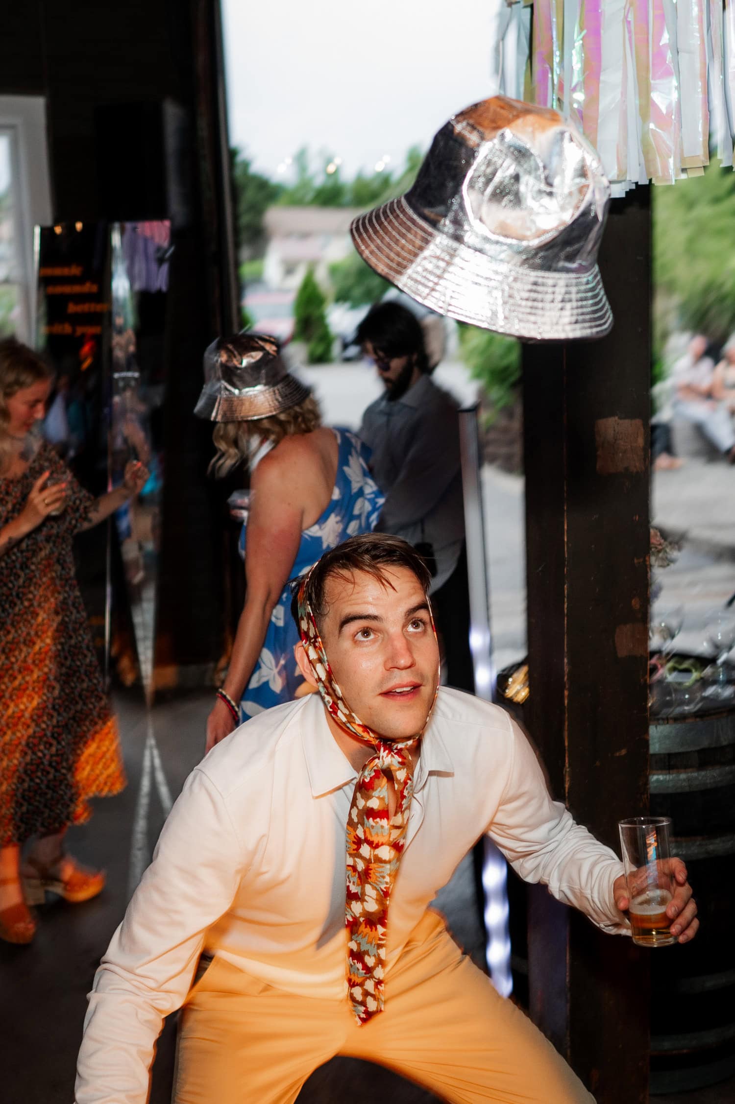A guest balances a hat on their nose while others in colorful clothing stand and laugh in the background at a Rossi Farms gathering.