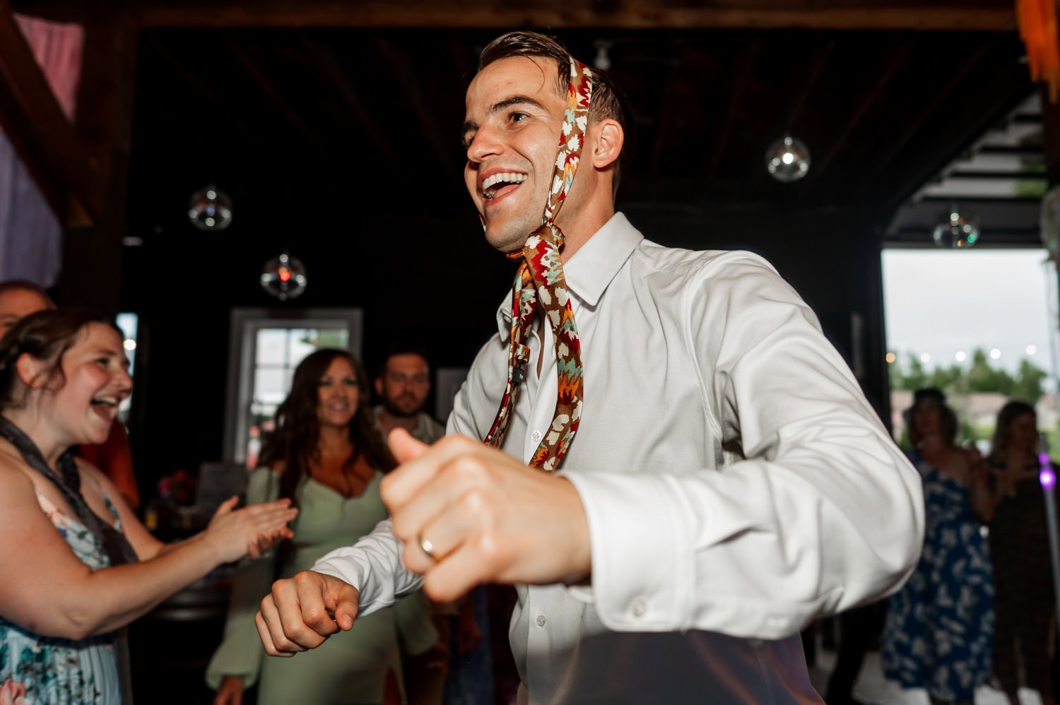 A man dancing with a tie on his head surrounded by smiling and clapping guests at Rossi Farms wedding.