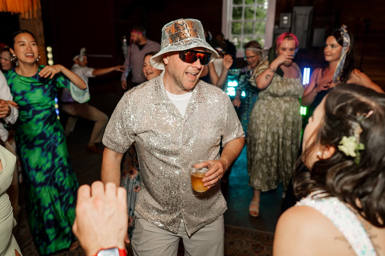 A man wearing a hat and sunglasses dances with a drink in hand at a lively celebration. Partygoers around him cheer and join in on the dance floor, all captured in a moment of joy and excitement.