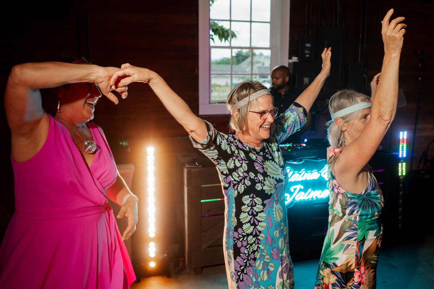 Three women wearing headbands dance joyfully under colorful lights at a lively indoor event.