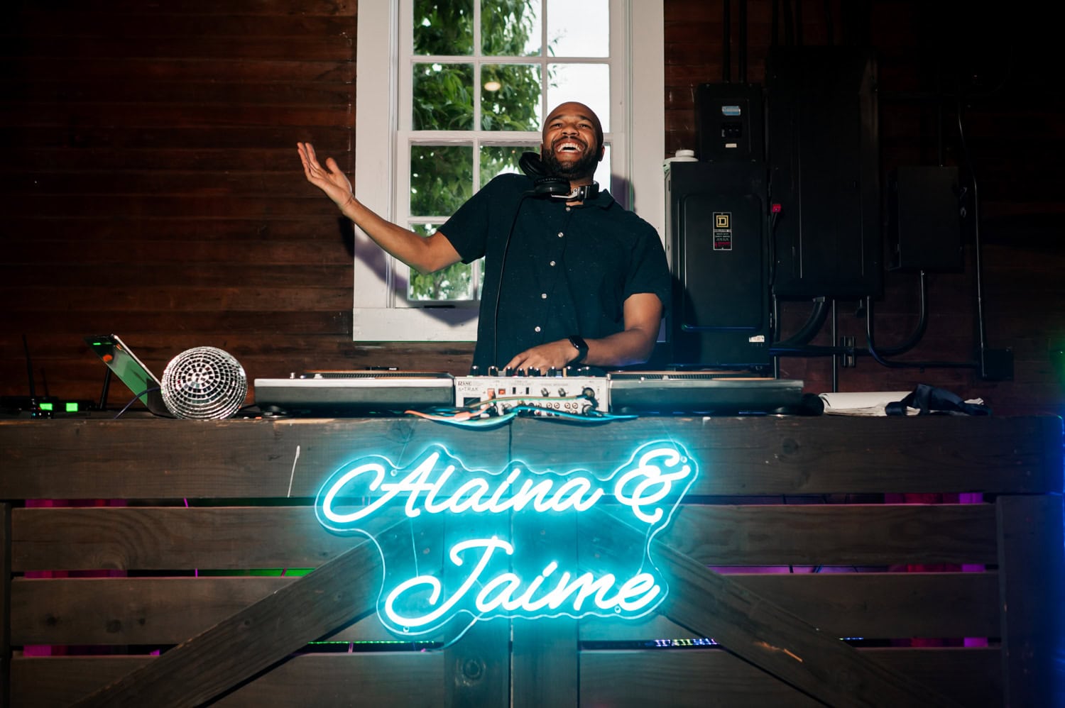 DJ smiling behind the booth with a neon sign that reads Alana & Jaime and a vintage microphone in front, at Rossi Farms wedding in a rustic setting.