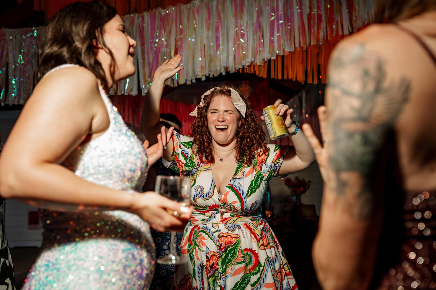 A lively party scene where a group of people dance with joy. One individual, wearing a colorful dress, holds a can and stands out among the crowd. The atmosphere is full of energy and celebration.