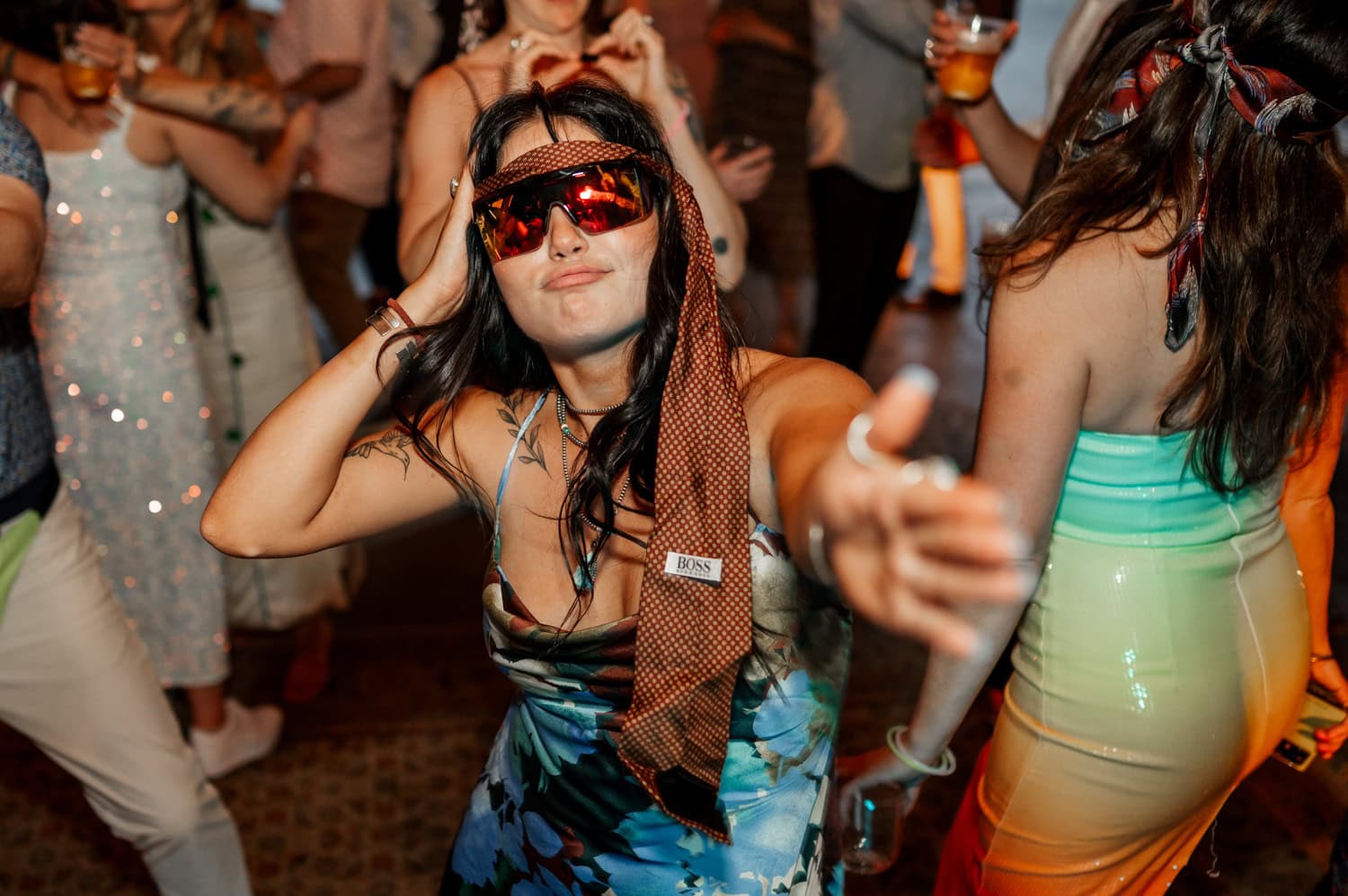 A woman in a blue dress and tie, wearing sunglasses, dances joyfully at a lively party with guests in colorful outfits.