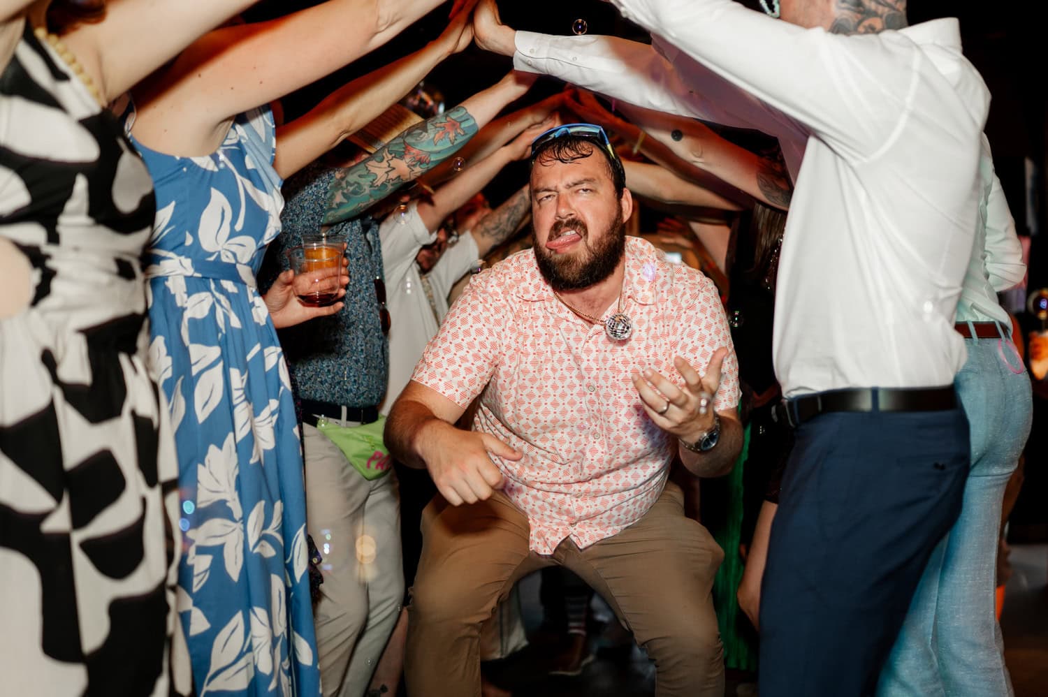 A man crouches and laughs under an arch of people with raised arms at a festive event, as seen in the Rossi Farms wedding photos.