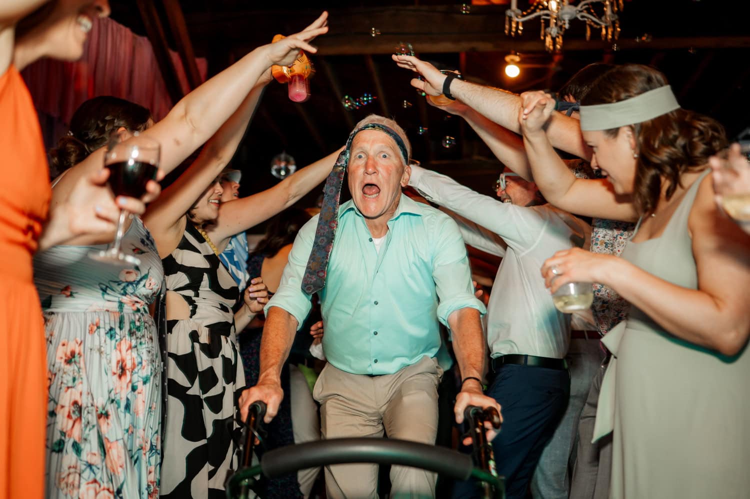 A man using a walker joyfully moves through a cheering crowd, dressed in festive clothes under an arch formed by raised arms. The atmosphere is lively and celebratory.