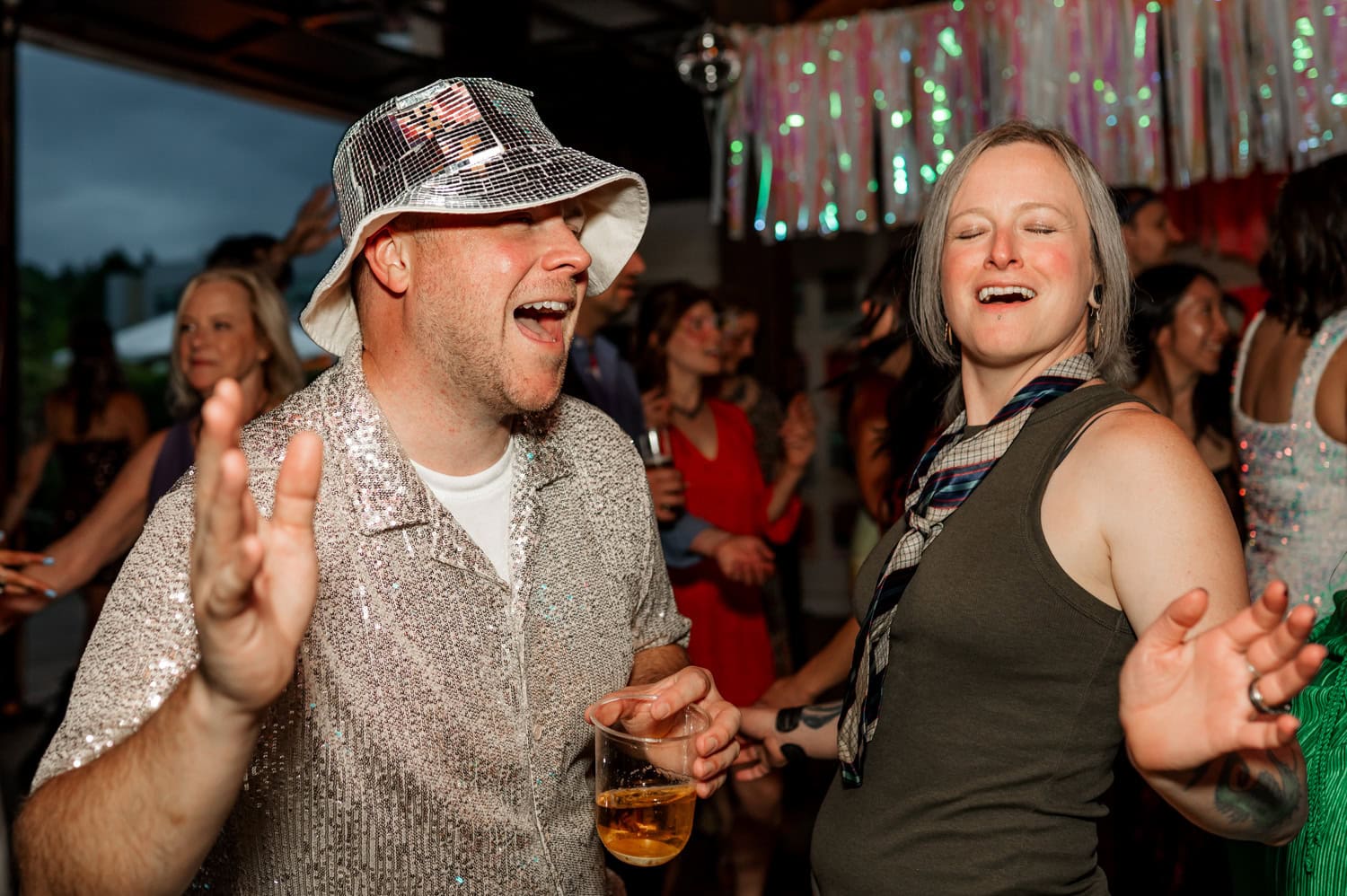 People dance energetically at a lively celebration. One partygoer stands out wearing a shiny bucket hat and shirt, holding a drink. Others in colorful outfits dance joyfully around them.