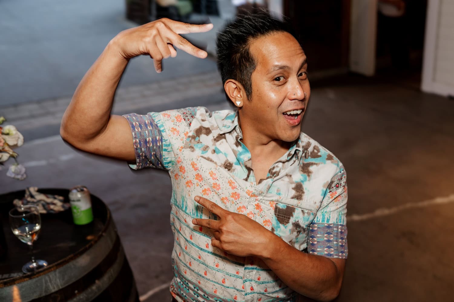 A person in a floral shirt playfully poses with hand gestures next to a wooden table with a drink.