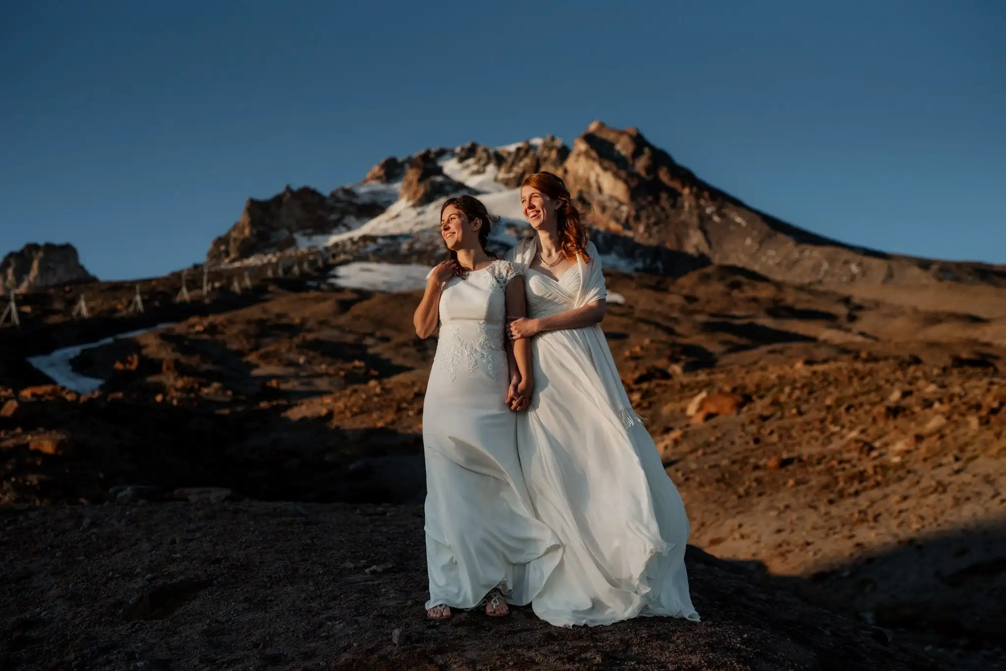 Amidst a rocky landscape with majestic mountains as their backdrop, two brides in white dresses stand hand in hand at sunset, captured beautifully by a Portland wedding and elopement photographer.