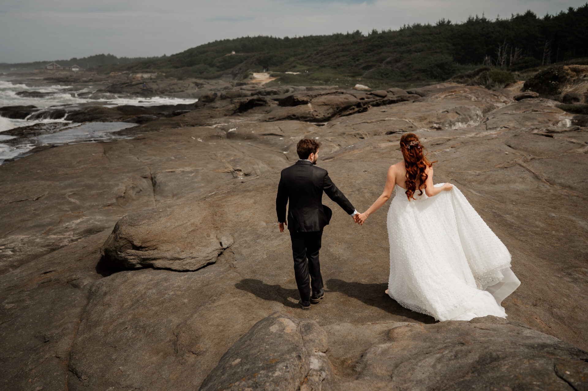 The bride and groom walk hand in hand across a rocky coastal landscape with a cloudy sky above, taking wedding photos at the Yachats Inn.