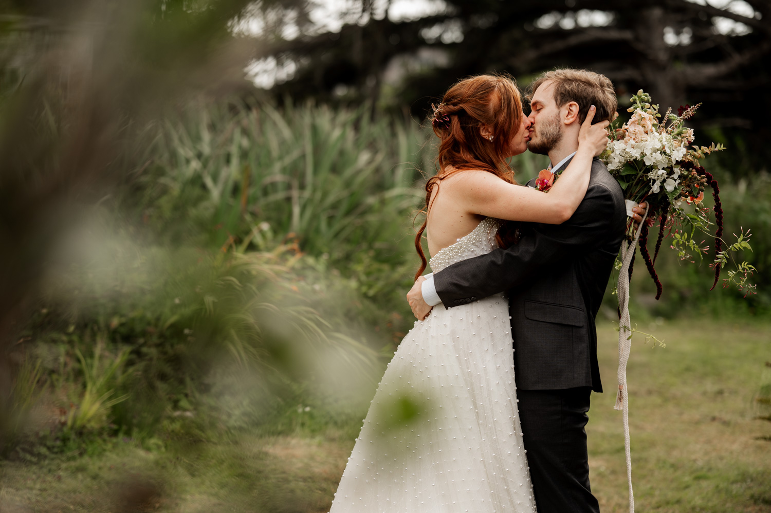 A bride and groom share a tender kiss outdoors surrounded by lush greenery, with the bride holding a bouquet. Theyre capturing this moment for their wedding photos at the Yachats Inn.