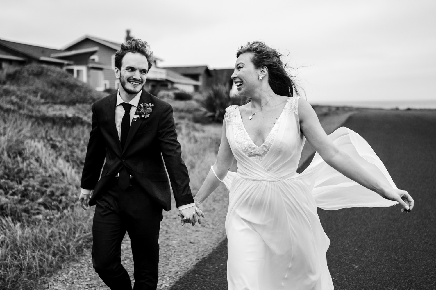 A happy couple in wedding attire joyfully runs hand in hand on a country road near the Yachats Inn with charming houses visible in the background.